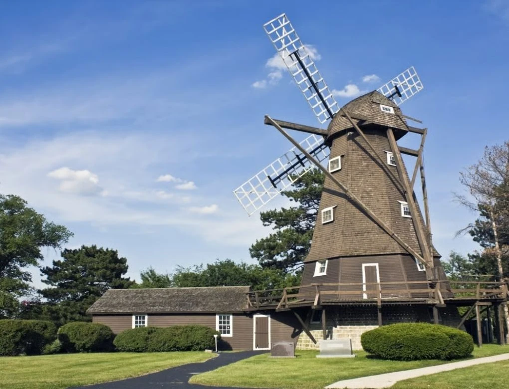 A historic, rustic wooden windmill and outbuilding surrounded by trees and a paved walking path.