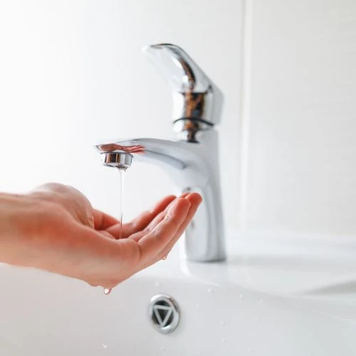 A person checking a very weak trickle of water flowing from a modern chrome bathroom sink faucet due to low water pressure.