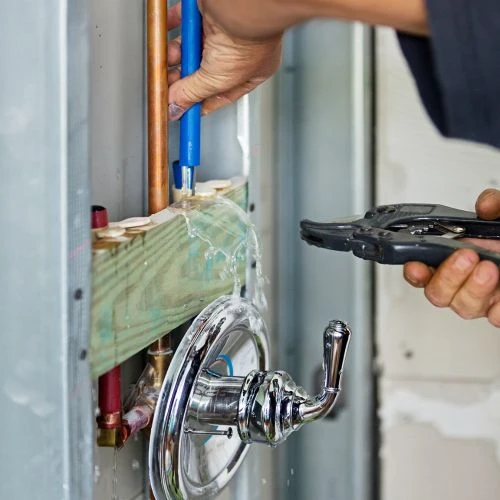 Close-up of a plumbing technician cutting blue and red PEX water lines next to a newly installed chrome shower valve assembly.