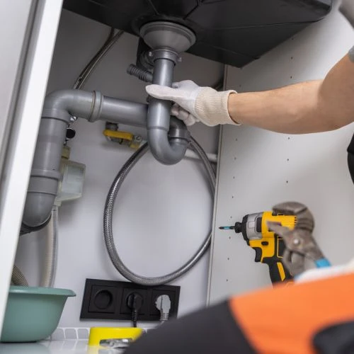 Close-up of a plumbing technician wearing protective gloves repairing and adjusting a grey PVC drain P-trap underneath a kitchen sink unit.