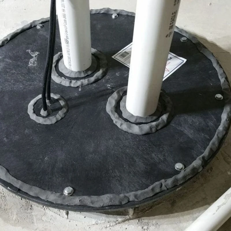 Close-up of a professionally sealed sewage ejector pump basin cover with dual PVC pipes and gas-tight caulking to prevent basement sewage odors in Northwest Chicago.