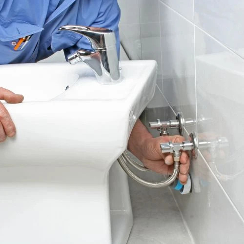 Close-up of an installer connecting flexible metal water lines and shut-off valves to a newly fitted bathroom porcelain bidet fixture.