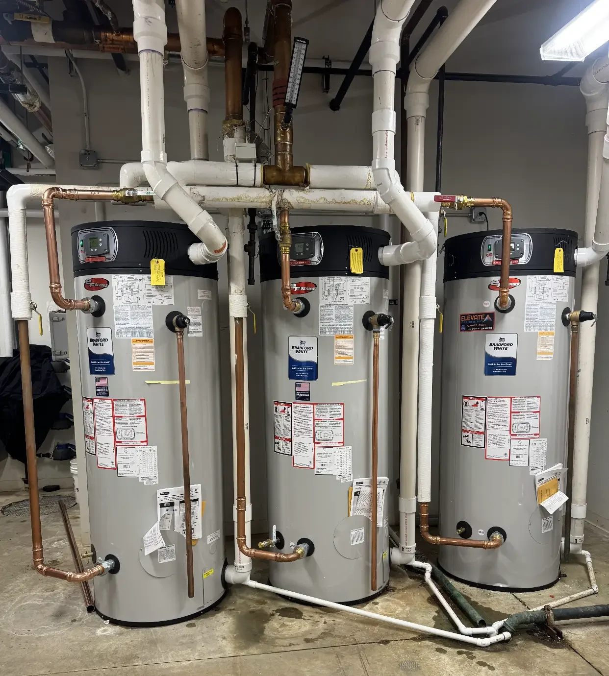 Commercial water heater installation by Elevate Plumbing featuring three high-capacity Bradford White units connected with complex copper and PVC piping.