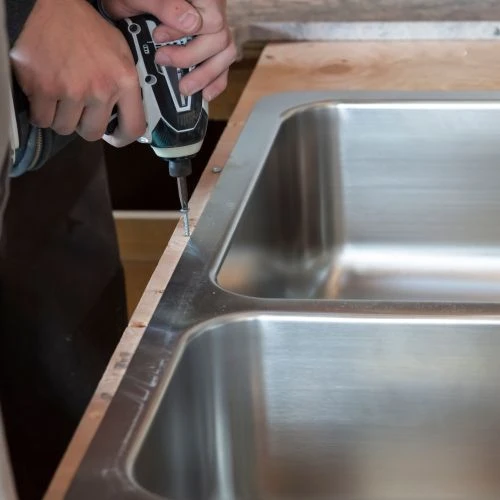 Contractor using a cordless electric drill to secure the rim of a stainless steel double sink to a wooden countertop base during installation.