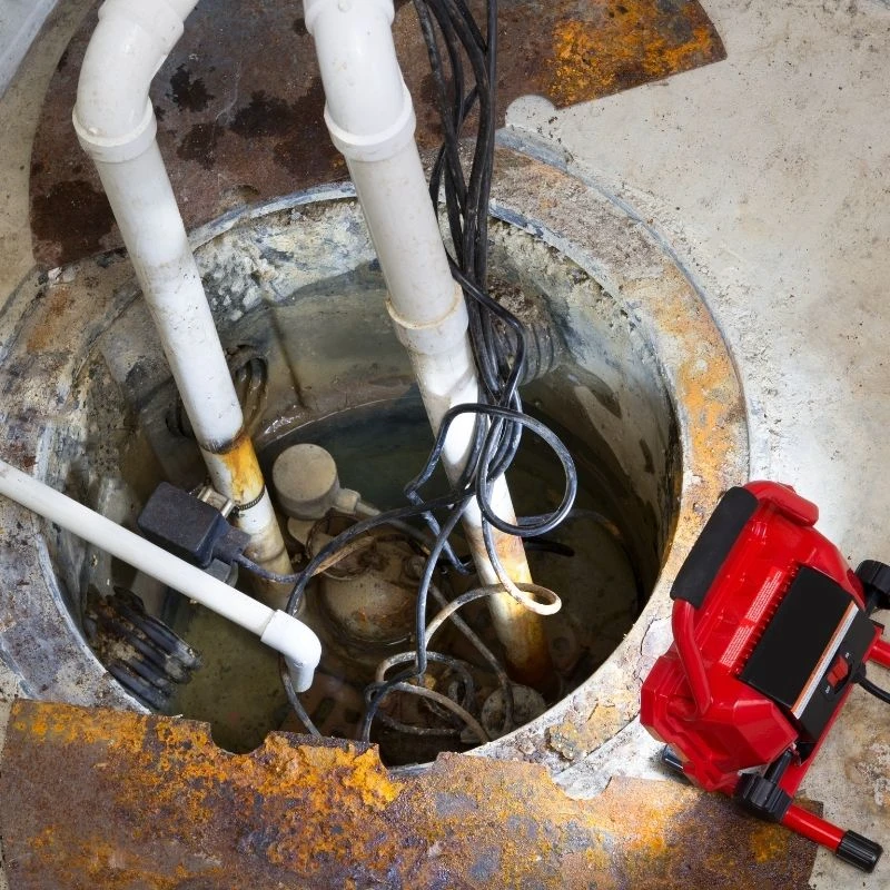 Elevate Plumbing work light illuminating an aging, tangled sump pump system with rusted pipes in need of a complete replacement in a Niles home.