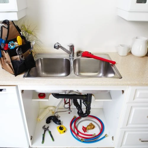 Kitchen sink installation area showing plumbing tools, a pipe wrench, red and blue supply lines, and an open layout setup inside an under-sink cabinet.