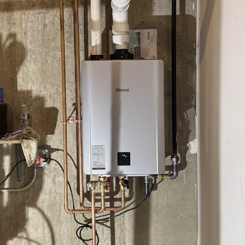 Newly installed silver Rinnai tankless water heater with expertly crafted copper piping and proper PVC venting, wall-mounted against a concrete wall.