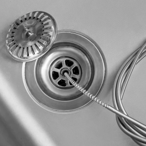 Overhead view of a flexible metal plumbing snake cable being fed down into a stainless steel kitchen sink drain to clear a stubborn clog.