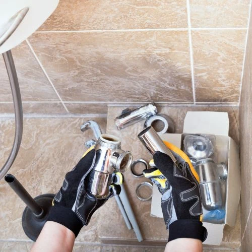 Overhead view of a worker wearing safety gloves assembling a new polished chrome sink drain trap alongside new replacement plumbing parts.