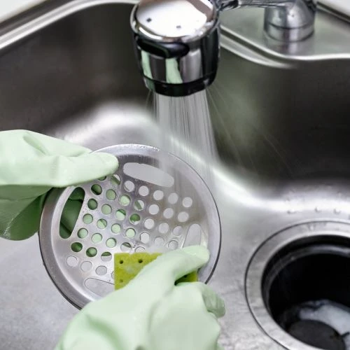 Person wearing green rubber safety gloves using a sponge to clean a stainless steel kitchen sink basket strainer under running water.