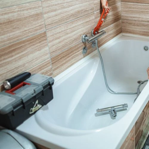 Plumber using a red pipe wrench to install a modern chrome bathtub faucet mixer valve on a tiled bathroom wall.