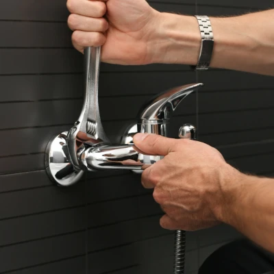 Plumber using an adjustable crescent wrench to tighten a chrome wall-mounted faucet fixture against a dark tiled bathroom wall.