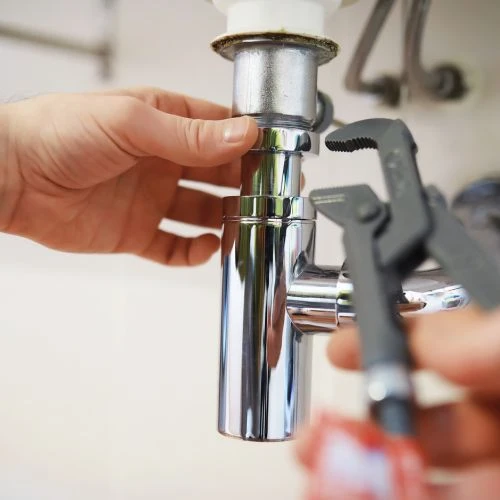 Plumbing technician using slip-joint pliers to secure a shiny chrome P-trap drain assembly underneath a bathroom washbasin.