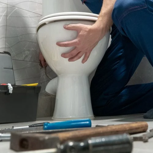 Side view of a professional plumber installing a new white ceramic toilet bowl base onto a tiled bathroom floor.