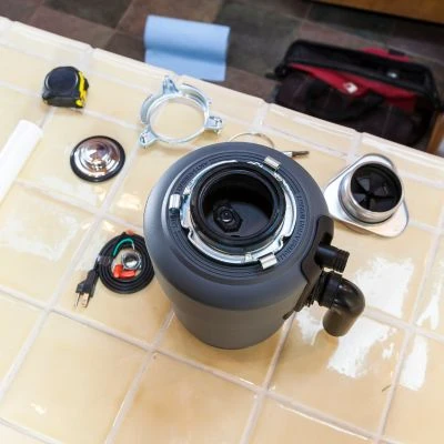 Top-down view of a brand new garbage disposal unit laid out on a kitchen tile floor alongside its mounting hardware, sink flange, and power cord parts.
