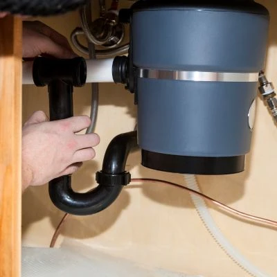 Under-sink view of a technician connecting black PVC drain pipes and a P-trap assembly to a garbage disposal unit.